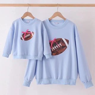 A matching mother-and-child sweatshirt set in light blue, hanging on wooden hangers from a wooden rod. The larger adult sweatshirt features a brown American football graphic with a pink bow, while the smaller child sweatshirt features a smaller matching football graphic.