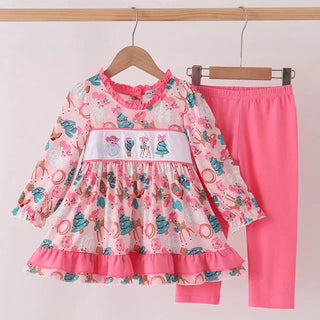 A toddler girl's two-piece clothing set hanging on wooden hangers against a plain light background. The set includes a long-sleeve smocked dress with a pink and multicolor floral print, a smocked white waistband with embroidered bows, and solid pink leggings.