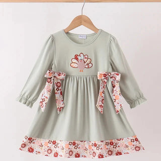 A toddler's long-sleeve dress in sage green, hanging on a wooden hanger. The dress features a floral patterned ruffle hem, matching floral ribbon bows on the sides, and a turkey embroidery on the chest.