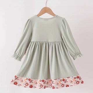 A light sage green long-sleeve dress for toddlers, hanging on a wooden hanger. The dress features a gathered waist and a contrasting ruffled hem with a red, pink, and orange floral pattern on a white background.