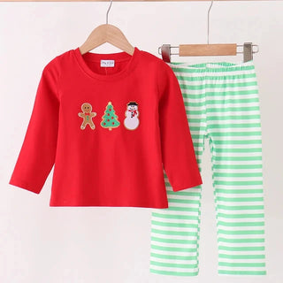 A children's two-piece holiday pants set hanging on wooden hangers against a plain light background. The set includes a bright red long-sleeve top with gingerbread man, Christmas tree, and snowman graphics on the chest, and light green and white horizontally striped pants.