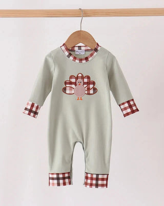 A baby long-sleeve romper in sage green, featuring a brown and white gingham pattern on the collar, cuffs, and hem, with a brown and white gingham turkey graphic on the chest. The romper is hanging on a wooden hanger from a wooden clothes rack against a plain off-white background.
