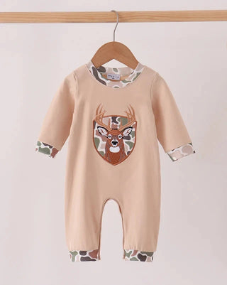 A baby long-sleeve romper in a light peach-beige color, hanging on a wooden hanger. The romper features a large embroidered deer head graphic on the chest with camouflage accents, and matching camouflage trim on the cuffs and leg openings.
