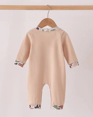 A baby long-sleeve footless romper in a light peach-beige color, featuring a subtle camouflage pattern trim at the collar and cuffs, hanging on a wooden hanger against a light beige background.