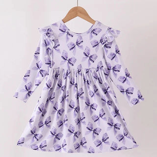 A long-sleeve girls dress with a purple bow pattern on a white background, hanging on a wooden hanger from a light wood rod against a plain off-white wall.