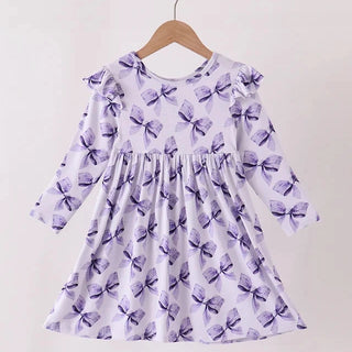 A long-sleeve girls dress with a purple bow pattern on a white background, hanging on a wooden hanger from a wooden rod against a plain light background.