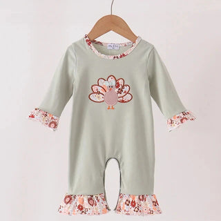 A baby long-sleeve romper in sage green, featuring a turkey graphic on the chest and floral ruffle trim at the cuffs and hem, hanging on a wooden hanger against a plain white wall.