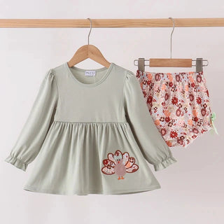 A toddler girl's two-piece outfit set hanging on wooden hangers from a clothing rack. The set includes a sage green long-sleeve tunic dress with a turkey graphic on the front, and matching floral bloomers with a red, pink, and orange pattern on a white background.