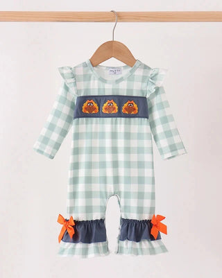 A baby girl's long-sleeve romper in a light green and white gingham pattern, featuring a navy blue chest band with three embroidered orange chicks, a navy blue ruffled hem with orange ribbon bows, and ruffled shoulder straps. The romper is shown hanging on a wooden hanger against a plain off-white background.