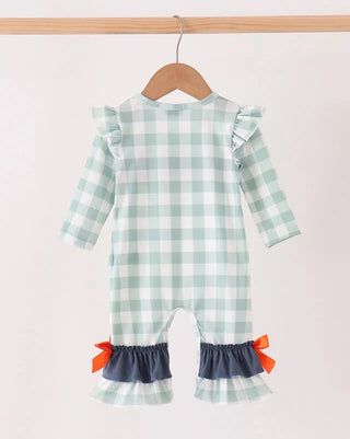 A baby girl's long-sleeve romper in a light mint green and white gingham check pattern, featuring ruffled shoulder details, a navy blue ruffled hem, and small orange bows, hanging on a wooden hanger.
