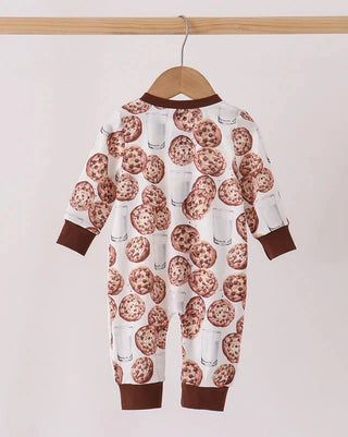 A baby long-sleeve footless one-piece jumpsuit with an all-over chocolate chip cookie and milk glass print, hanging on a wooden hanger from a light wood rod against a plain off-white background.