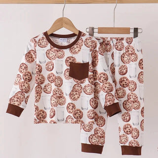 A two-piece children's pajama set hanging on wooden hangers against a plain light background. The set includes a long-sleeve top and matching pants with an all-over chocolate chip cookie print on a white base, plus solid brown trim on the collar, sleeve cuffs, pocket, and pant cuffs.