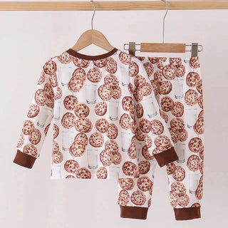 A two-piece children's pajama set hanging on wooden hangers against a plain light background. The set includes a long-sleeve top and matching pants with an all-over print of chocolate chip cookies and milk glasses on a white base, finished with solid dark brown cuffs and collar trim.
