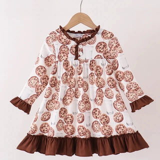 A toddler girl's long-sleeve dress with a white base printed with brown chocolate chip cookies and illustrated milk bottles, finished with a dark brown ruffled collar, cuffs, hem, and front tie detail, hanging on a wooden hanger.