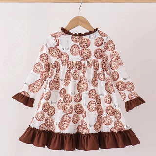 A long-sleeve baby dress with an all-over chocolate chip cookie print, featuring brown ruffle trim at the collar, cuffs, and hem, hanging on a wooden hanger against a plain light background.