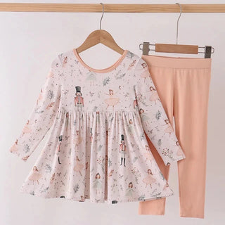 A children's two-piece outfit set hanging on a wooden hanger bar. The set includes a long-sleeve smocked dress in a light pink and white print with nutcracker motifs, and matching solid light peach-pink leggings.