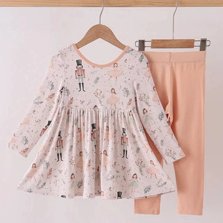 A two-piece toddler girl's outfit hanging on wooden hangers against a plain white wall. The set includes a long-sleeve tunic top in a light pinkish-beige fabric with a whimsical print of nutcrackers, fairies, and floral motifs, paired with solid peach-pink leggings.