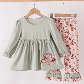 A two-piece baby girl's outfit set hanging on wooden hangers against a plain white wall. The set includes a sage green long-sleeve peplum top with a turkey patch on the front, and matching floral print pants in red, orange, and pink on a white background.