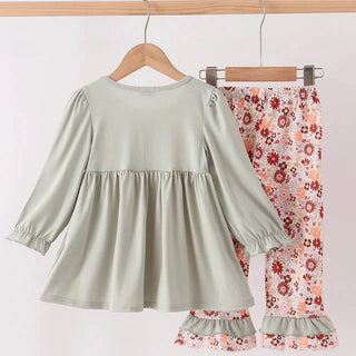 A two-piece children's outfit hanging on wooden hangers from a wooden rod. The set includes a sage green long-sleeve peplum-style top and matching floral print pants in pink, red, and white with ruffled hems.