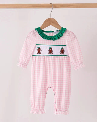 A baby long-sleeve romper in a pink and white gingham pattern, featuring a green ruffled collar, a white chest band with three embroidered gingerbread cookies, and ruffled leg hems. The romper is displayed on a wooden hanger against a plain light background.