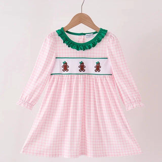 A pink and white gingham long-sleeve dress hanging on a wooden hanger against a light wall. The dress features a green ruffled collar, a white smocked chest panel with three gingerbread cookies, and elasticated cuffs.