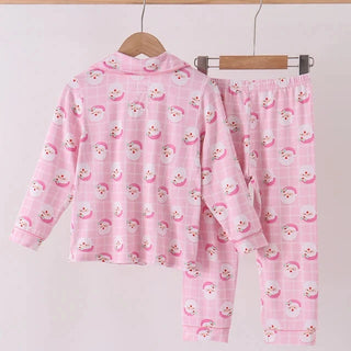 A two-piece children's pajama set in light pink, featuring a long-sleeve top and matching pants with a repeating cartoon character print, hanging on wooden hangers from a wooden rod against a plain light background.