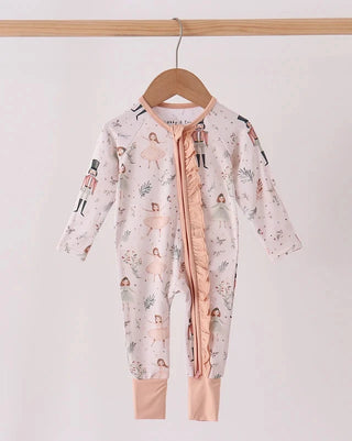 A baby long-sleeve footless romper hanging on a wooden hanger from a wooden rod. The romper is light pink with a whimsical print of ballerinas, nutcricks, and small flowers, featuring a peach-colored ruffled front zipper and matching peach cuffs.