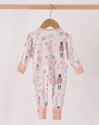 A baby long-sleeve footless jumpsuit hanging on a wooden hanger, featuring a light pink background with a whimsical print of nutcrackers, ballerinas, and tiny floral motifs, finished with peach-colored cuffs and collar.