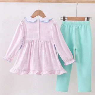 Pink checkered dress with white collar and green pants on hangers against a light gray background