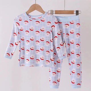 A two-piece children's pajama set hanging on wooden hangers against a plain light background. The set includes a long-sleeve top and matching pants in light blue with an allover print of red Santa hats on a subtle grid pattern.