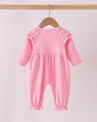 A pink baby jumpsuit with long sleeves, ruffled shoulder details, and elasticized leg cuffs, hanging on a wooden hanger from a light wood rod against a plain light background.