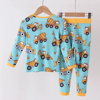 A two-piece children's pajama set hanging on wooden hangers against a plain white wall. The set includes a long-sleeve top and matching pants in light blue, covered in a playful print of yellow construction vehicles, with yellow trim at the collar, cuffs, and waistband.