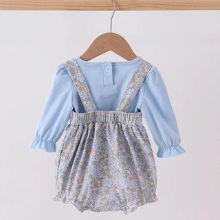 A baby girl's two-piece outfit hanging on a wooden hanger. The set includes a light blue long-sleeve top with ruffled cuffs and a matching light blue floral overall romper with patterned shoulder straps, hanging against a plain light background.