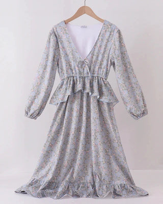 A long-sleeve V-neck maxi dress with a ruffled peplum waist, featuring a delicate multicolor floral pattern on a light gray-blue base. The dress is displayed on a wooden hanger hanging from a wooden clothes rack against a plain white wall.