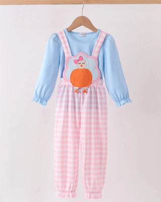 A baby girl's long-sleeve jumpsuit hanging on a wooden hanger. The jumpsuit features a light blue long-sleeve top layered under pink and white gingham check overalls, with a decorative applique of a cartoon turkey on the chest.