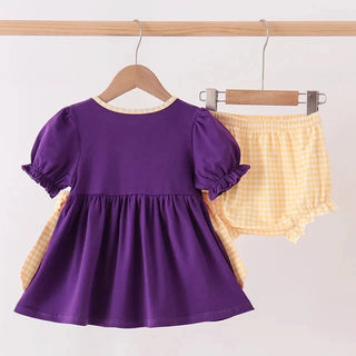 A two-piece toddler girl's outfit hanging on wooden hangers against a plain white wall. The set includes a purple short-sleeve puff-sleeve dress with yellow gingham side panels and matching yellow gingham shorts.