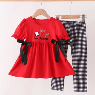 A toddler girl's two-piece outfit set hanging on wooden hangers against a plain white wall. The set includes a bright red short-sleeve dress with black ribbon bows on the sleeves and a 'Go Dawgs' graphic on the chest, paired with black and white gingham checkered pants.