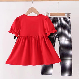 A toddler girl's two-piece outfit set hanging on wooden hangers against a white wall. The set includes a bright red short-sleeve peplum-style dress with ruffled sleeves and a small black bow on the side, paired with black and white gingham checkered pants.