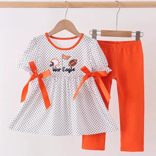 A toddler girl's two-piece outfit set hanging on wooden hangers against a plain white wall. The set includes a white short-sleeve top with black polka dots, orange trim, and two orange ribbon bows on the sleeves, paired with solid bright orange leggings.