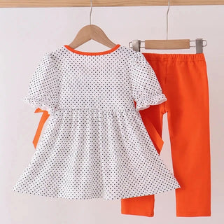 A toddler girl's two-piece outfit hanging on wooden hangers against a plain light background. The set includes a white short-sleeve dress with black polka dots and orange trim, paired with bright orange leggings.