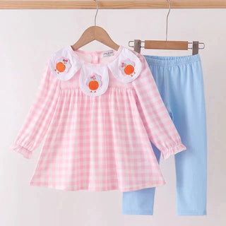A toddler girl's two-piece outfit set consisting of a pink and white gingham long-sleeve top with white Peter Pan collar and embroidered orange fruit details, paired with light blue leggings, hanging on wooden hangers against a white wall.