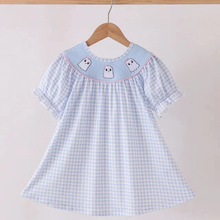 A girl's short-sleeve dress with a light blue and white gingham pattern, featuring a light blue collar decorated with three embroidered white ghosts and a thin pink border. The dress is shown hanging on a wooden hanger from a wooden clothes rack against a plain, light beige wall.