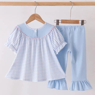 A two-piece girl's outfit set hanging on wooden hangers against a plain light background. The set includes a light blue and white gingham short-sleeve top with a solid light blue collar and pink piping, paired with matching light blue ruffled pants.