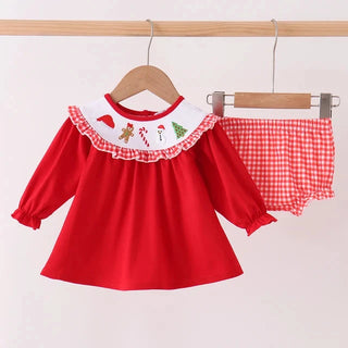 A toddler girl's two-piece holiday outfit hanging on wooden hangers against a plain white wall. The set includes a bright red long-sleeve top with a white ruffled collar decorated with small Christmas illustrations, and matching red-and-white gingham check bloomers.
