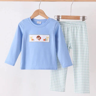 A children's two-piece clothing set hanging on wooden hangers against a plain white wall. The set includes a light blue long-sleeve top with a small cartoon bunny patch on the chest and light green and white gingham pants.