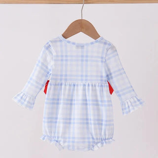 A baby girl's long-sleeve romper in a light blue and white gingham plaid pattern, hanging on a wooden hanger from a wooden clothes rack. The romper features ruffled cuffs, a ruffled hem, and small red side tabs.
