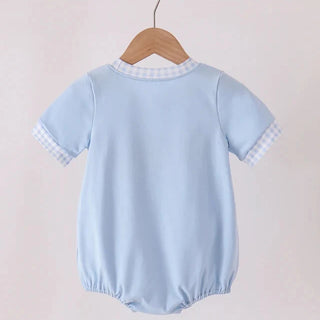 A light blue short-sleeve baby romper hanging on a wooden hanger from a wooden clothes rack, shown from the back against a soft, light pink background.