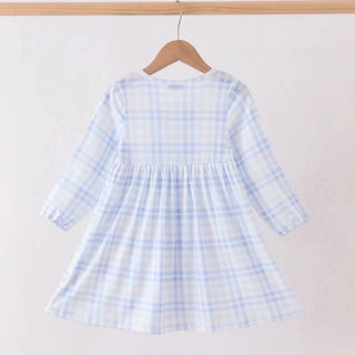A light blue and white plaid long-sleeve baby dress hanging on a wooden hanger from a wooden clothes rack against a plain off-white wall.