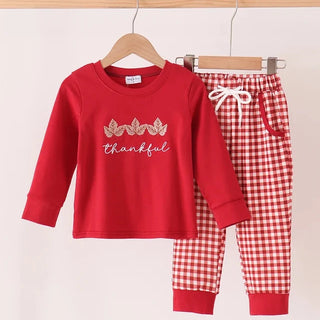 A toddler boy’s two-piece pajama set hanging on wooden hangers against a light beige background. The set includes a red long-sleeve top with a 'thankful' script and gold leaf design on the chest, and matching red and white gingham check pants with a white drawstring.