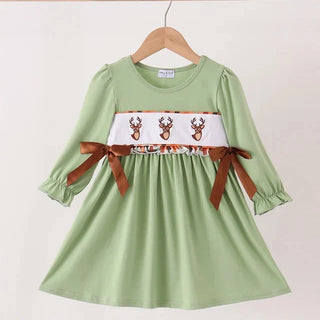 A light green long-sleeve dress for toddlers, hanging on a wooden hanger against a plain off-white wall. The dress features a white smocked chest panel decorated with three brown reindeer motifs and brown ribbon bows tied at the waist.